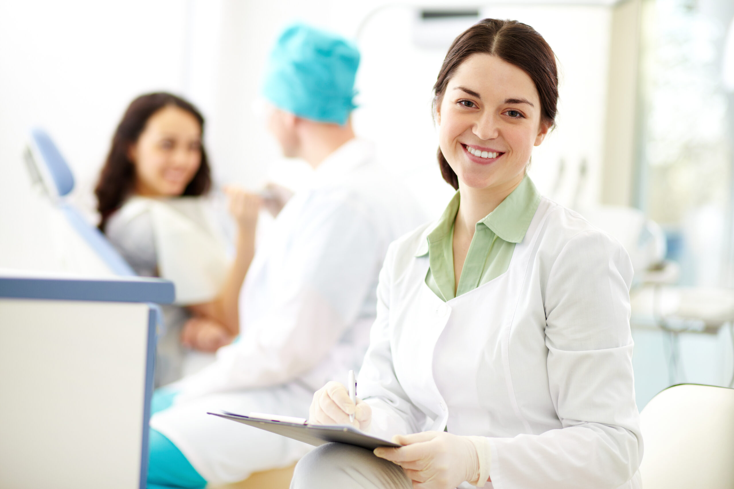 Pretty nurse looking at camera with smile on background of dentist and patient